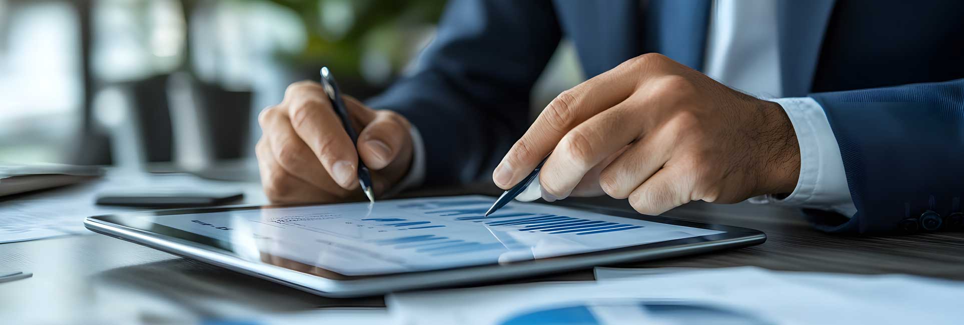 Close-up view of  hands analyzing data on a digital tablet, with documents and a laptop on an office desk.