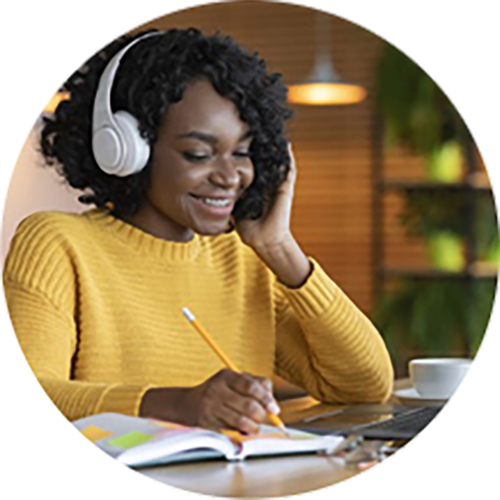  Student in gold sweater wearing headphones, taking online class with an instructor while writing notes.