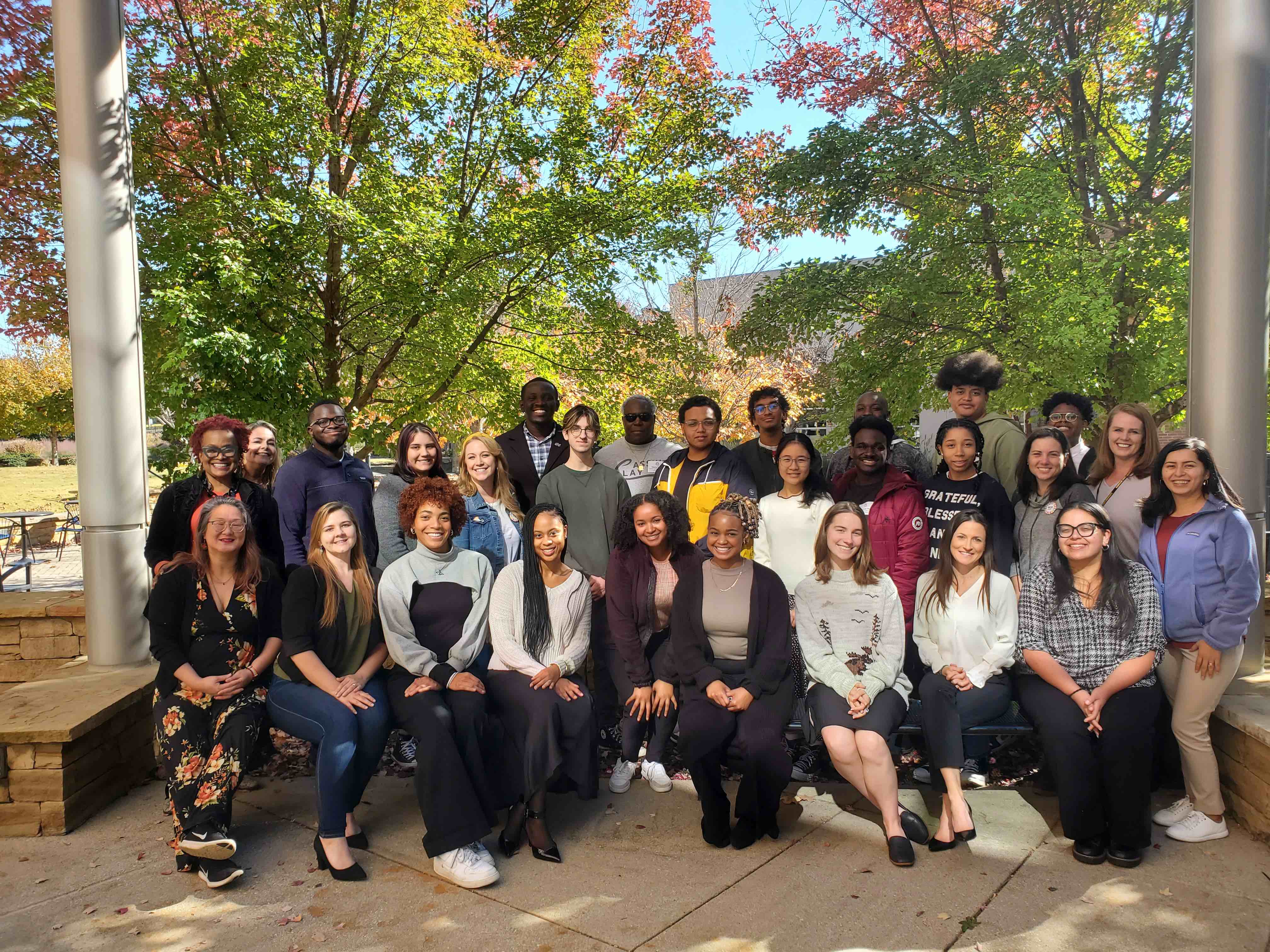 A diverse group of college students and adults stand together outside and smile