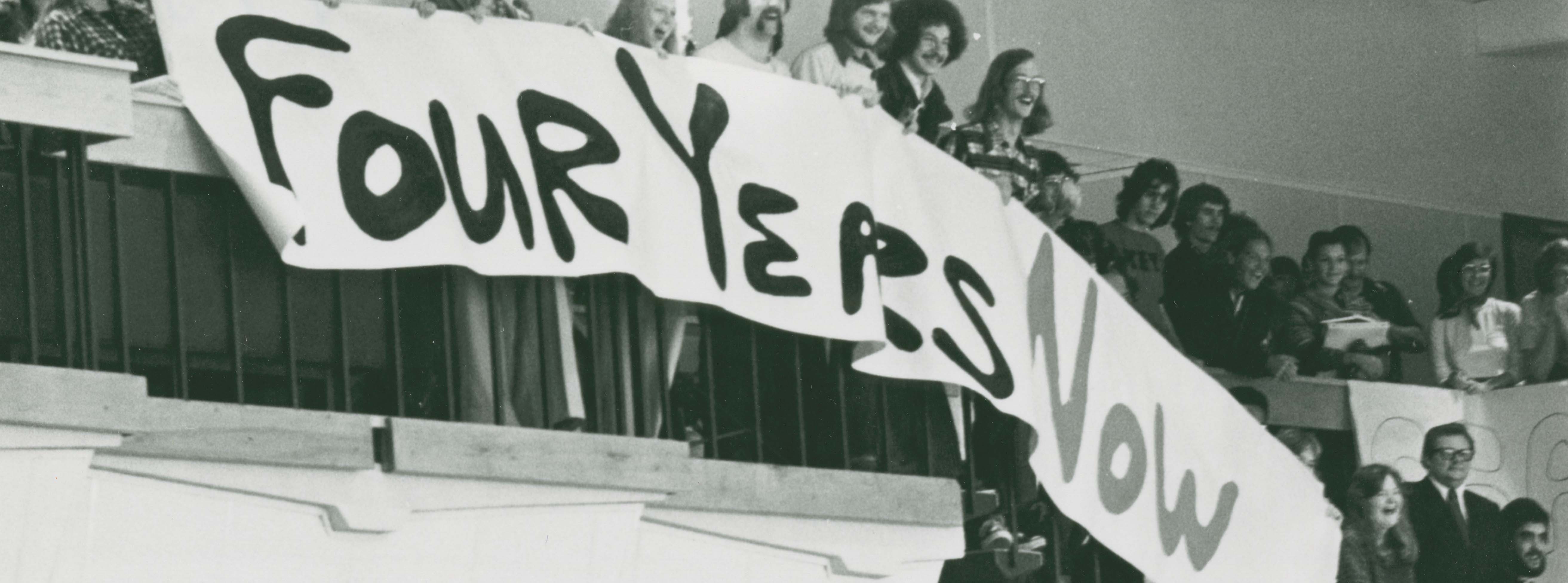 Students standing from a balcony unfurling a banner with the words Four Years Now written on it.