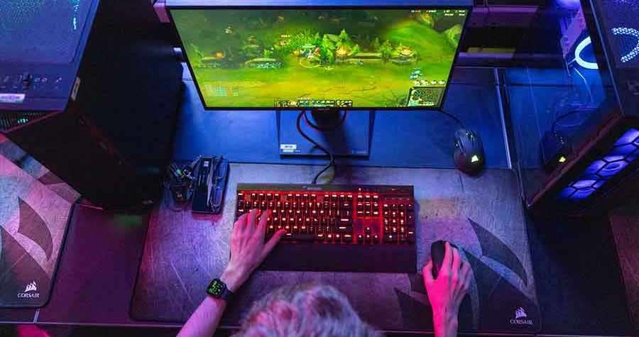 A person's hands typing on a red backlit keyboard with a gaming mouse next to it. A computer monitor displays a colorful gaming scene in the background