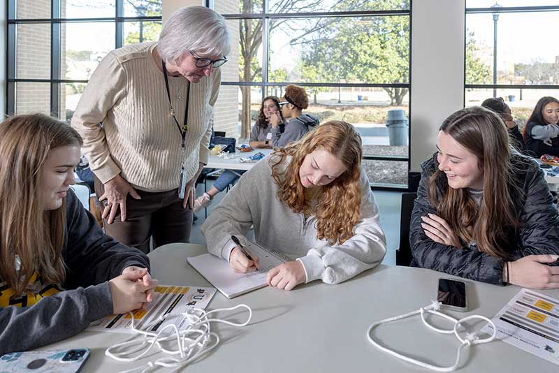 a class group around a table with a professor helping the students, with supportive communities for researchers across disciplines, promoting knowledge exchange, and enabling access to internal and external funding.